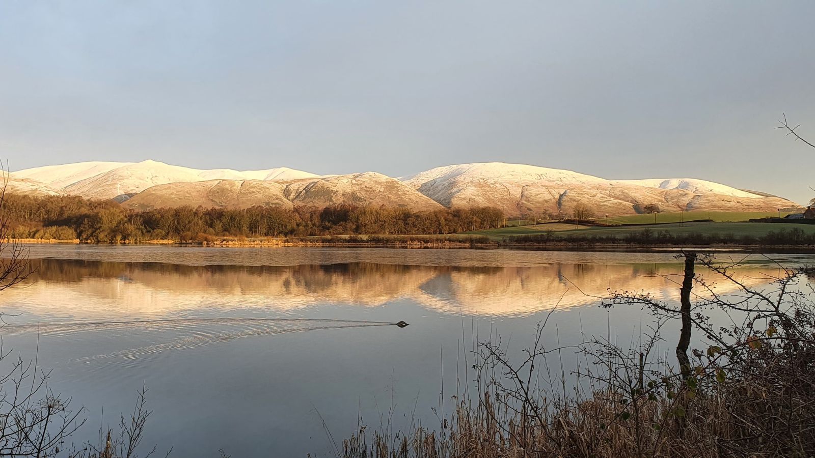 View across the water at Gartmorn Dam. The water is flat calm and the hills in the background have a light dusting of snow.